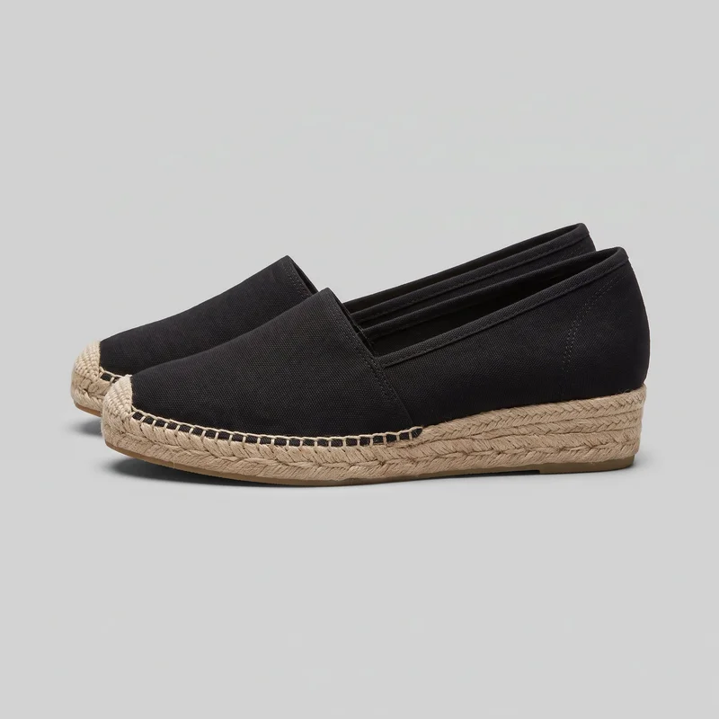 A pair of women's black slip-on espadrille platform flats is shown in a side/front angled view. The shoes have rounded closed toes, low-cut openings, loafer-like vamps, stitched upper seams, braided rope-wrapped platform midsoles, and thin flat outsoles.