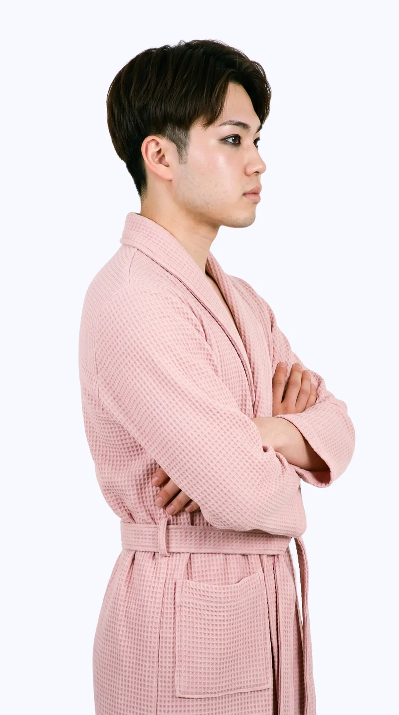 A 22-year-old Japanese man wears a blush pink robe in a three-quarter-length side view, standing with his arms crossed and a natural expression. A flat porcelain-colored backdrop frames the studio cutout.