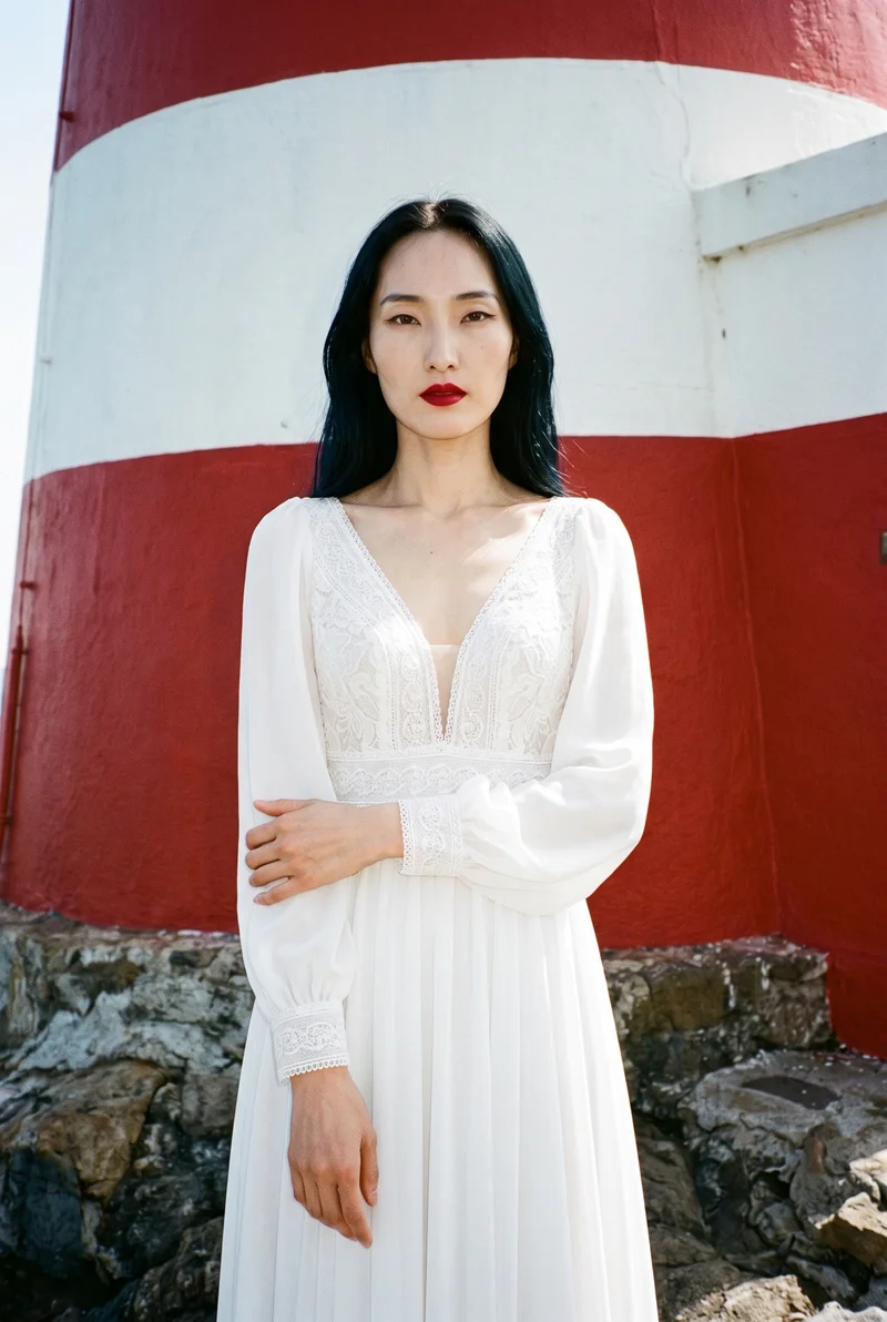 A 34-year-old Korean woman is shown from the upper body in a front view, wearing a bohemian chiffon wedding dress. She has a natural facial expression and is touching her wrist. A New England lighthouse forms the backdrop.