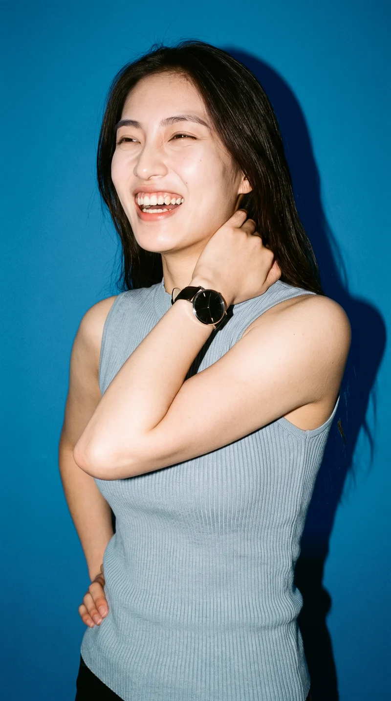 A 24-year-old Chinese woman is shown from the upper body at a front three-quarter angle, laughing with one hand behind her neck. She wears a women's black leather-strap analogue watch. A cerulean studio-wall backdrop fills the background.