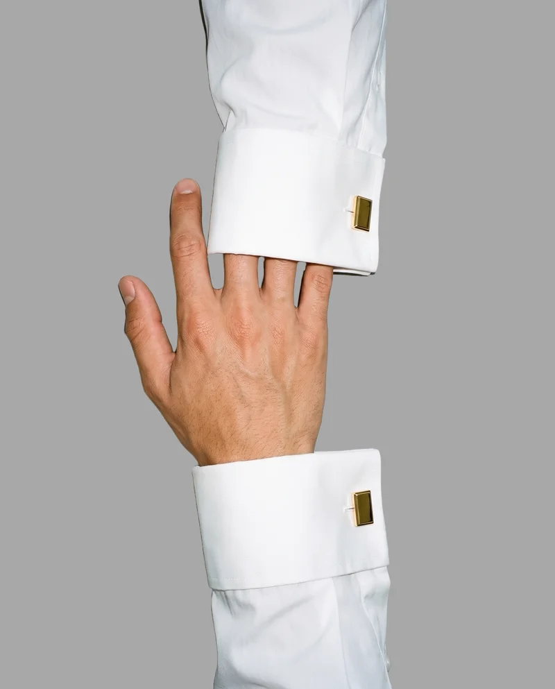 Gold classic cufflinks shown with a hand and wrist at rest in a top-view composition. The image is framed to focus on the hand and wrist area against a flat Pebble Grey backdrop.