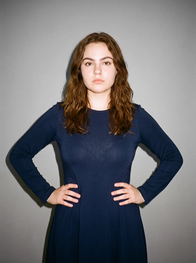 A 20-year-old Caucasian woman wears a navy long-sleeve modest dress in an upper-body front view, standing with her hands on her hips and an editorial expression. A pebble-grey studio-wall backdrop frames the composition.