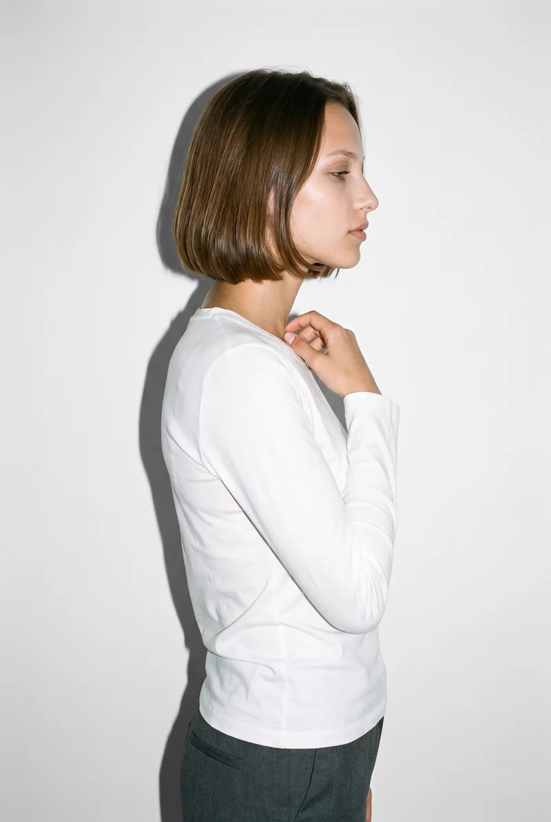 A 20-year-old Slavic woman is shown from the upper body in side view wearing a white long-sleeve tee. One hand rests at her collarbone, and she has a soft facial expression. A Seasalt studio-wall backdrop frames the composition.