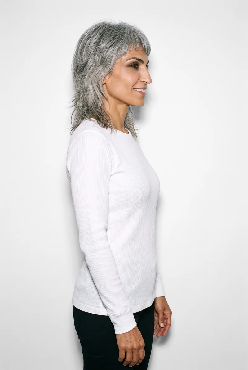 A 42-year-old Arab woman models a white women's base-layer thermal top in an upper-body side view, with her arms relaxed and a smile. The product is shown against a pure white studio-wall backdrop.