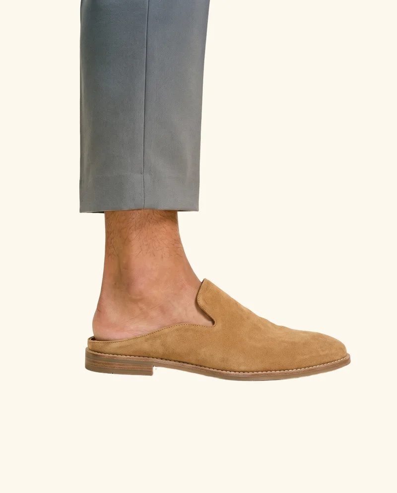A tan suede mule for men is shown from the side with the toe pointed. The shoe is framed at the ankle against a creamy white flat-color backdrop.
