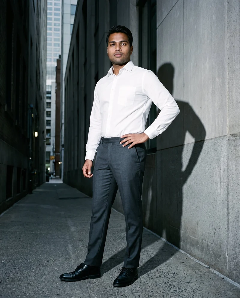 A 20-year-old Indian man wears charcoal suit trousers in a full-body angled-front view, standing with one hand on his hip. The Financial District urban canyon forms the backdrop.