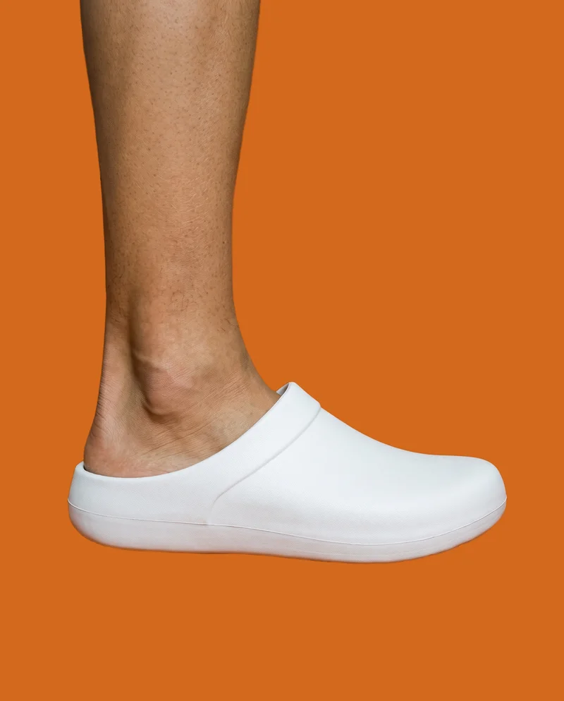 White men's clogs shown from the side at ankle level, framed from the ankle down against a flat cinnamon-colored backdrop.