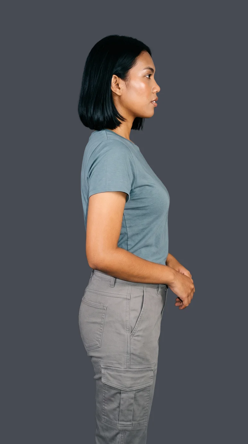 A 20-year-old Southeast Asian woman models grey cargo pants in a three-quarter-length side view, with her hands positioned in front and a neutral expression. The garment is shown against a flat graphite backdrop.