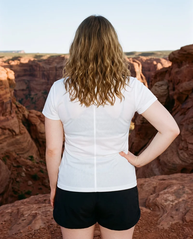 A 20-year-old Slavic woman is shown from the rear in an upper-body view, wearing a white mesh performance top with one hand on her hip. Red rock formations form the backdrop.
