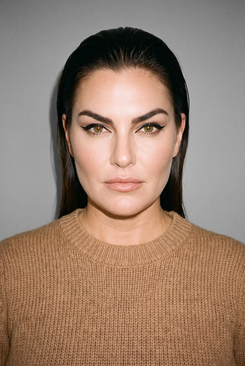 A 50-year-old Caucasian woman wears a women's camel knit pullover jumper in a bust-length front view. Her head is level, with an editorial facial expression, against a pebble grey studio-wall backdrop.