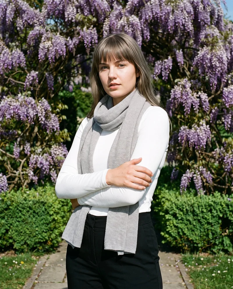 A 22-year-old Caucasian woman wears a grey wool scarf in an upper-body, angled-front view with her arms crossed and a sultry expression. A wisteria archway forms the backdrop.