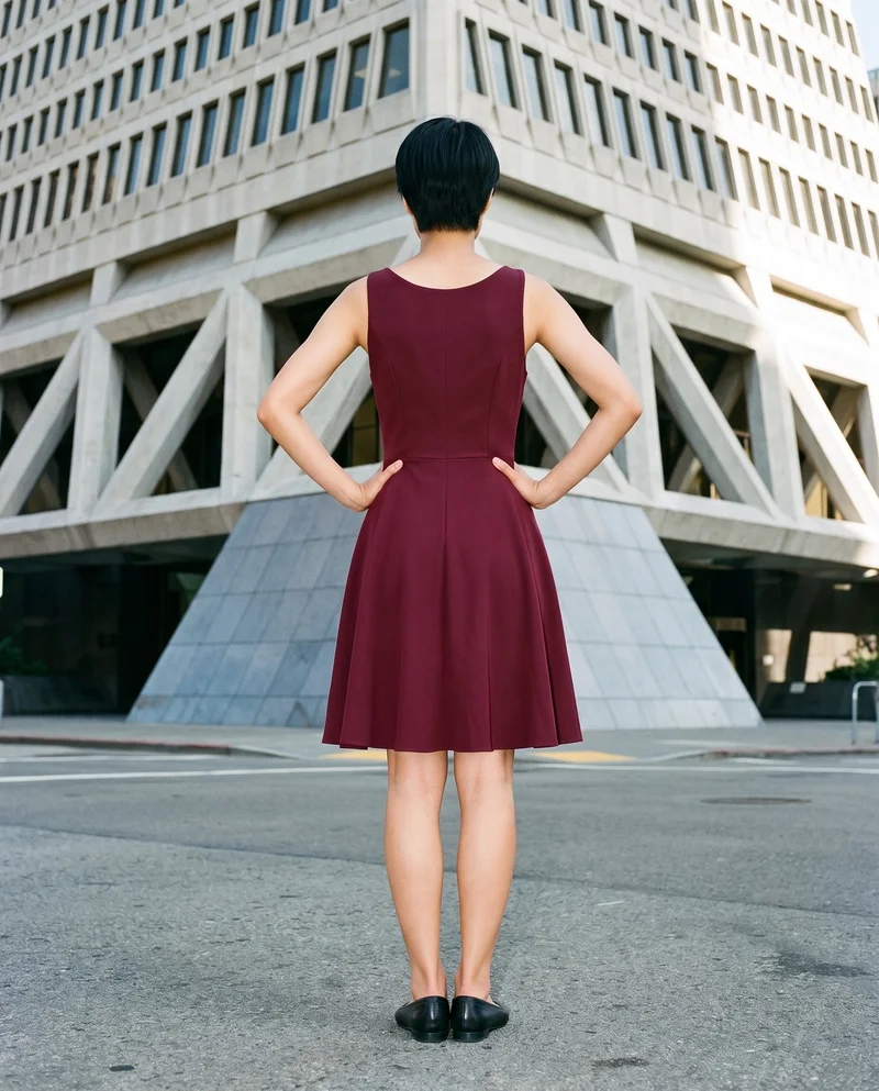 A 19-year-old Japanese woman models a burgundy cocktail dress in a full-body rear view, standing with her hands on her hips. A pyramid building appears as a low-angle backdrop.