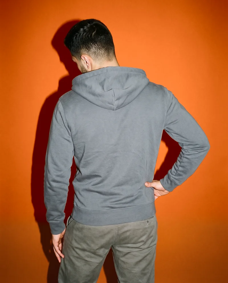 A 19-year-old Arab male model wears a charcoal cotton zip-up hoodie, shown from the back in a three-quarter-length frame with one hip angled outward. A burnt orange studio-wall backdrop fills the background.