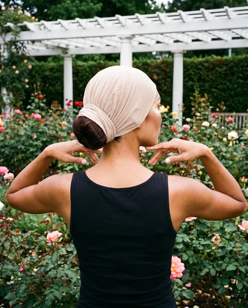 A 25-year-old Southeast Asian woman wears a beige cotton underscarf, shown in a head-only back view with her hands at her jaw. A rose garden pergola serves as the backdrop.