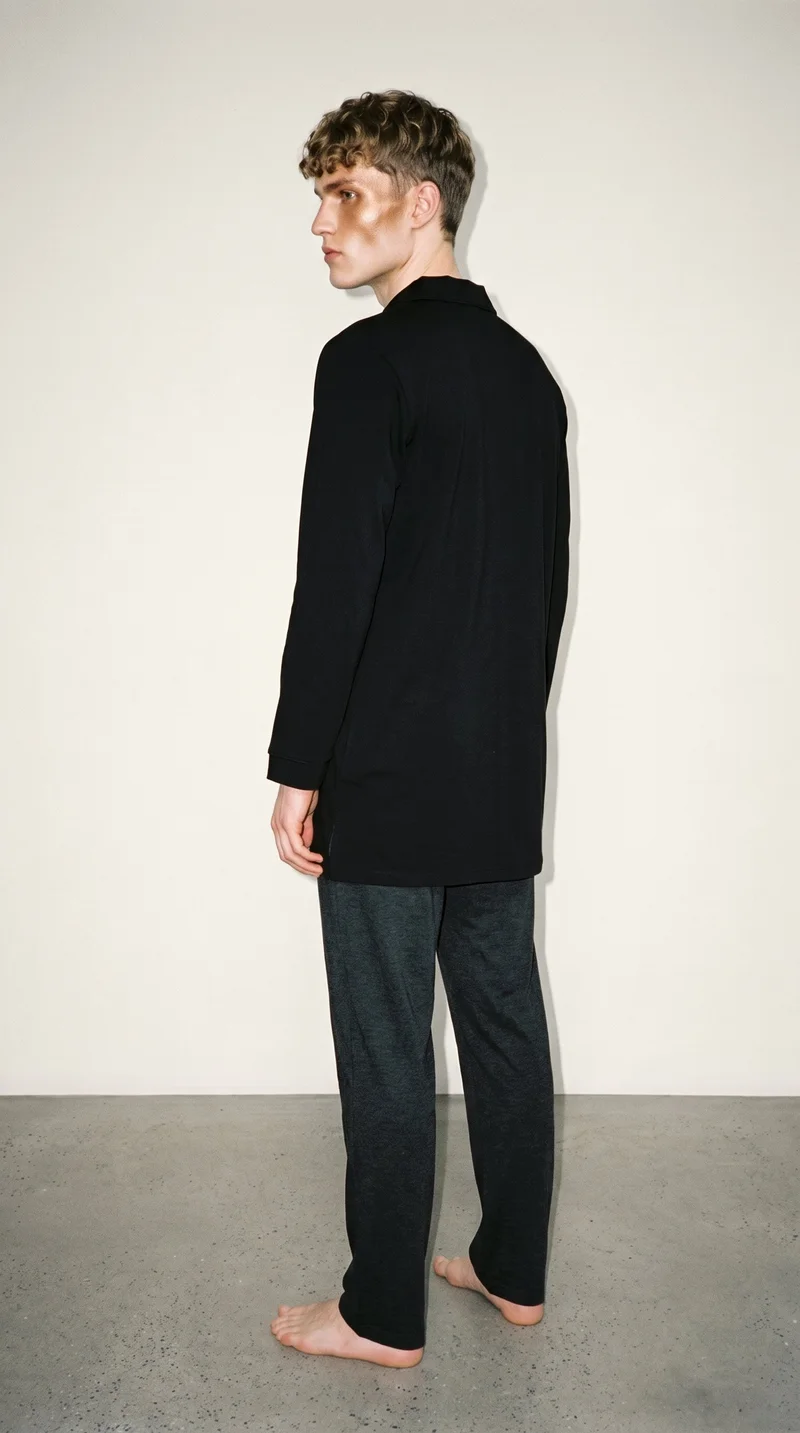 A 19-year-old Caucasian male model stands straight in a full-body, angled rear view wearing a black nightshirt. An Oyster Pearl studio-wall backdrop is visible behind him.