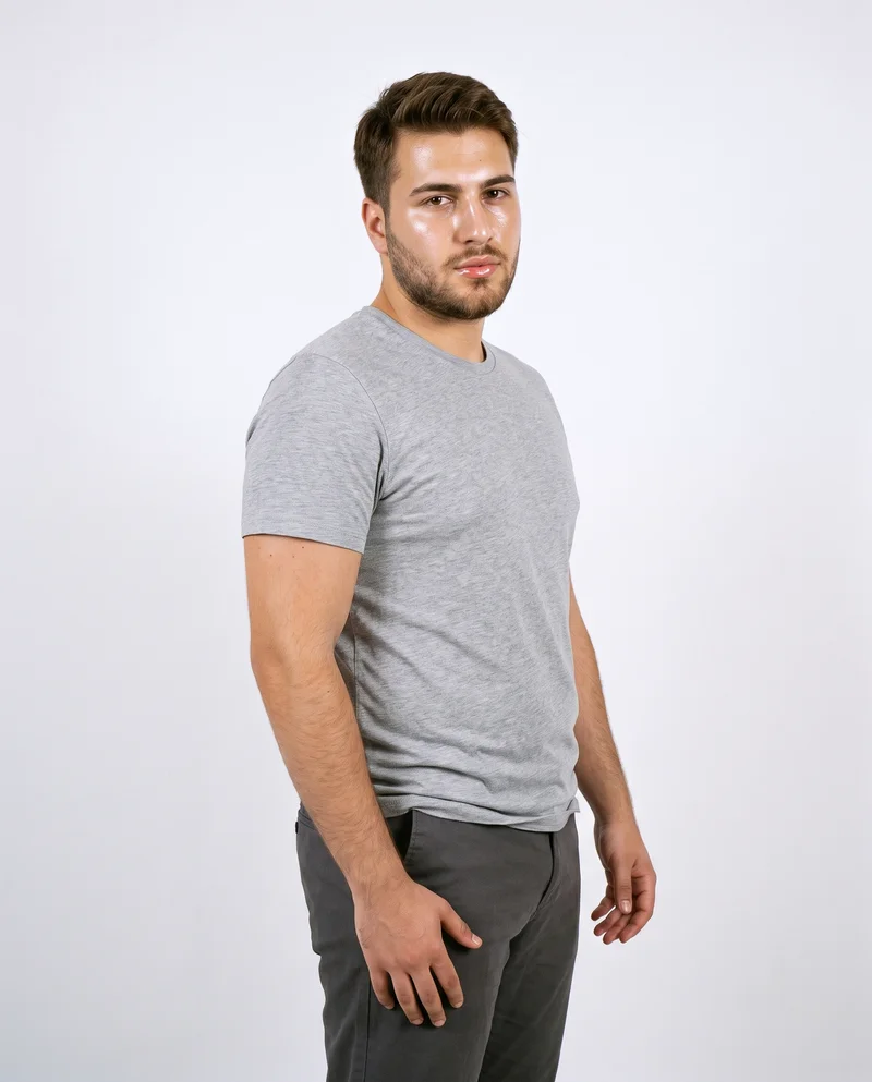 A 20-year-old Turkish male model wears a men's grey marl T-shirt against a porcelain studio-wall backdrop. He is shown from the front at a three-quarter angle in a three-quarter-length frame, with a waist-twist pose and a serious expression.