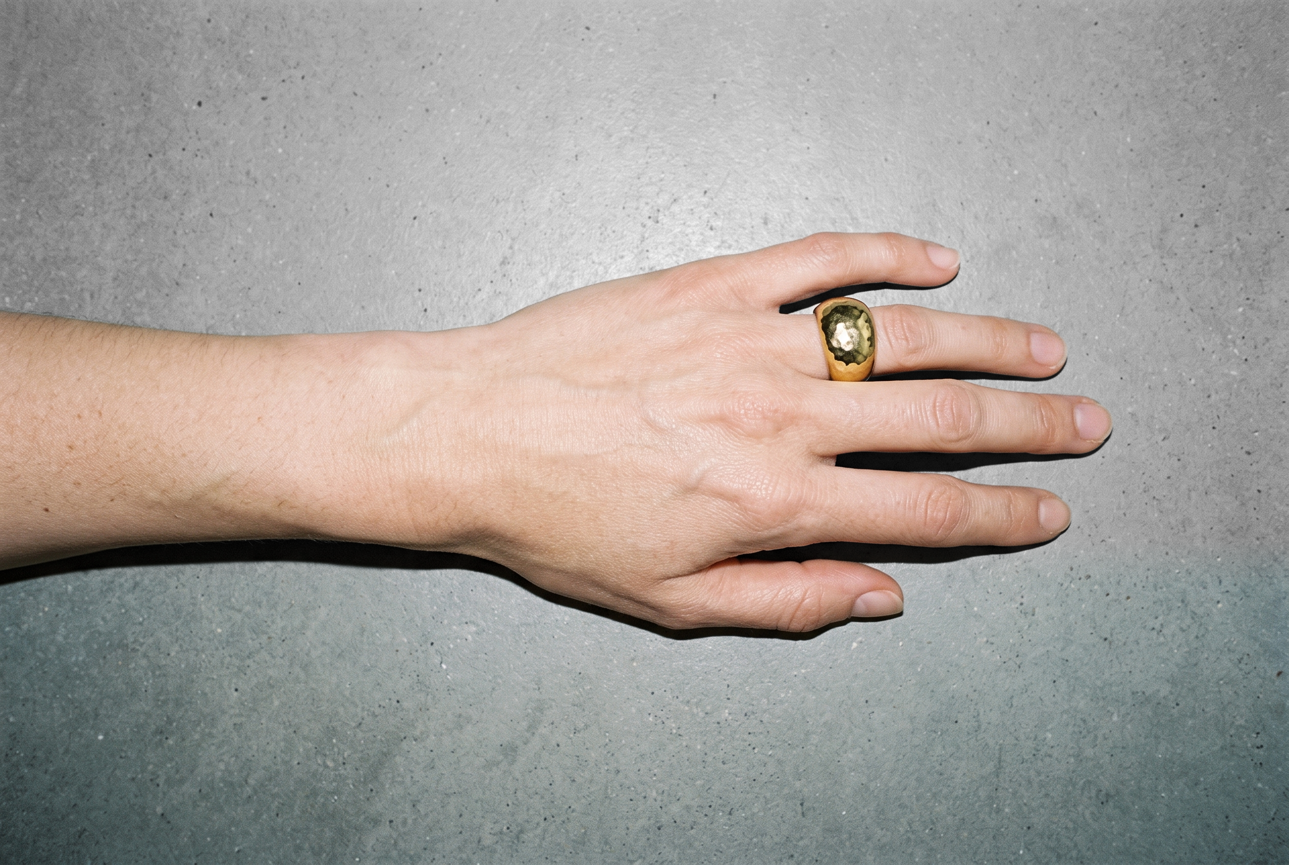 A chunky gold ring worn on a hand shown from above, with the hand and wrist resting against a pebble grey studio backdrop.