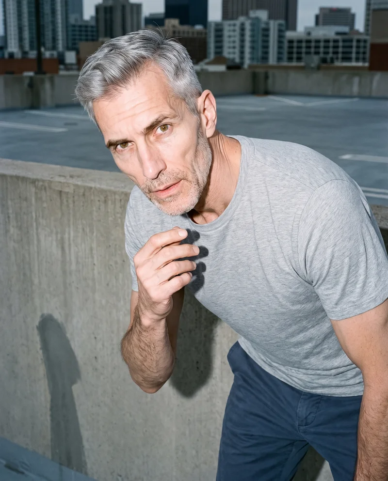 A 56-year-old Caucasian man wears a grey marl T-shirt in an upper-body view from a front three-quarter angle. One hand is held at his chin, and the backdrop is a parking garage rooftop.