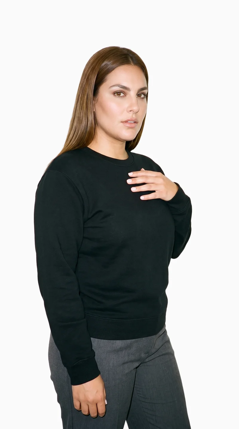 A 31-year-old Hispanic woman is shown from the upper body at a front three-quarter angle, wearing a black crewneck sweatshirt with her fingers at her chest. Her natural facial expression is visible against a flat Seasalt-colored backdrop.