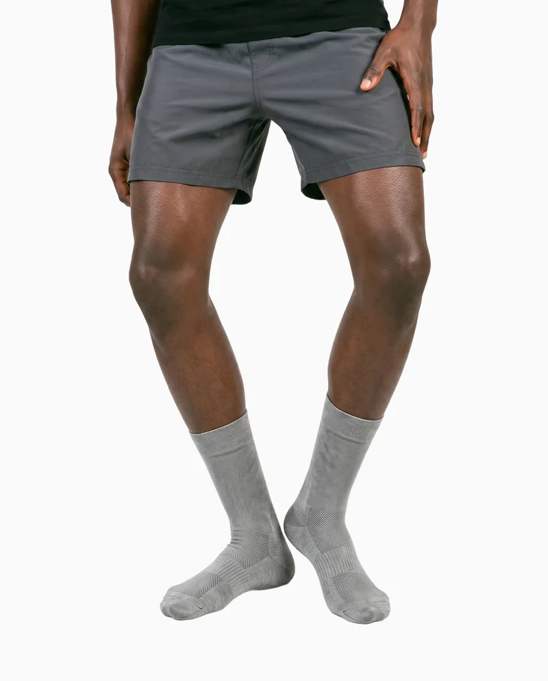 A male model's lower legs are shown from the front with the knees angled inward, wearing grey performance socks. The image uses a flat seasalt-colored backdrop.