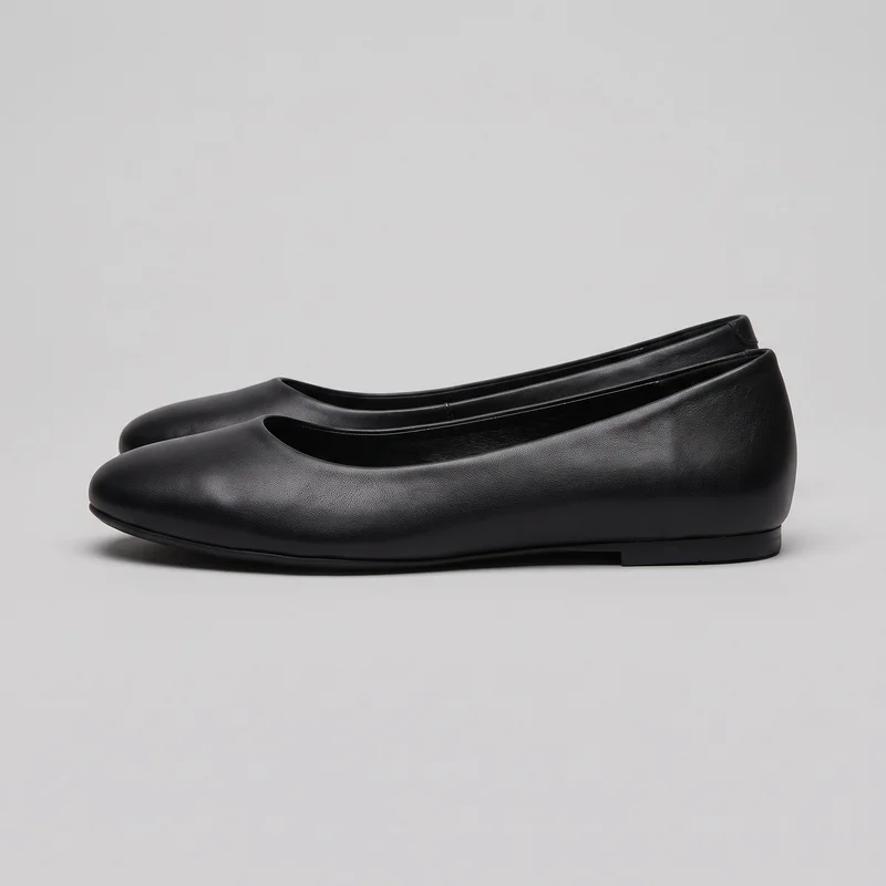 A pair of black women's ballet flats is shown in a side-oriented standalone view. The shoes have a low-profile slip-on shape, rounded almond toes, open toplines, closed heel counters, flat soles, and low stacked heel blocks. Their unadorned uppers have a fine pebbled texture, narrow raised binding around the toplines, and subtle seams along the sole edges.