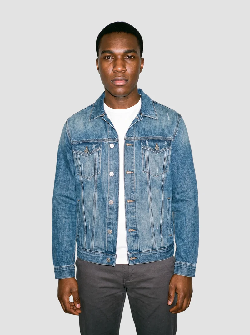 A 20-year-old African male model wears a men's mid-wash oversized denim jacket in a front-facing, three-quarter-length view. He stands in a relaxed pose with a neutral expression against a flat Fog Grey backdrop.