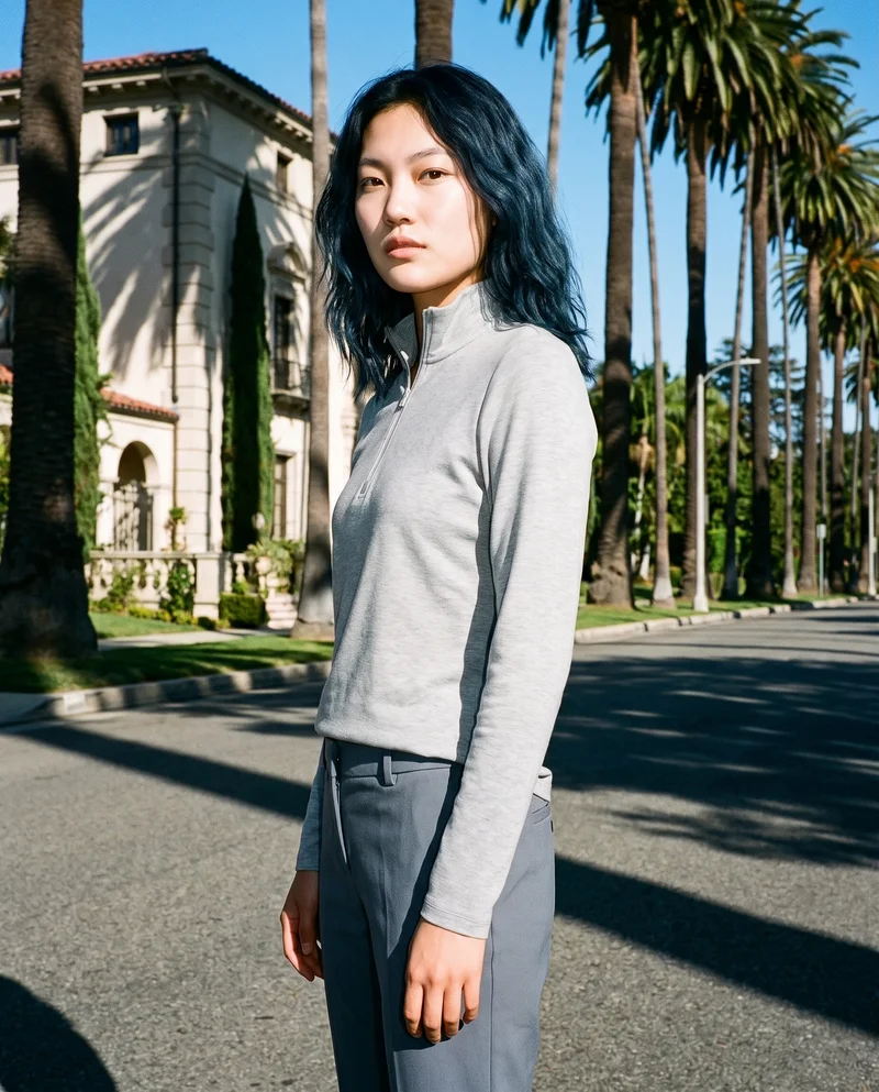 A 24-year-old Chinese woman wears a grey quarter-zip and stands in a three-quarter-length front view with one hand on her waist. Her serious expression is visible, with the Palm Mansion Boulevard backdrop behind her.
