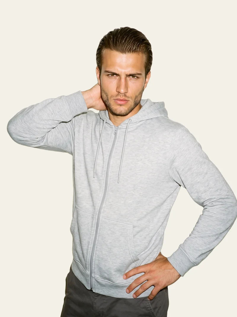 A 21-year-old Turkish male model wears a grey tracksuit top in an upper-body view from the front three-quarter angle. His hand is behind his neck and his expression is intense, against an alabaster flat-color backdrop.