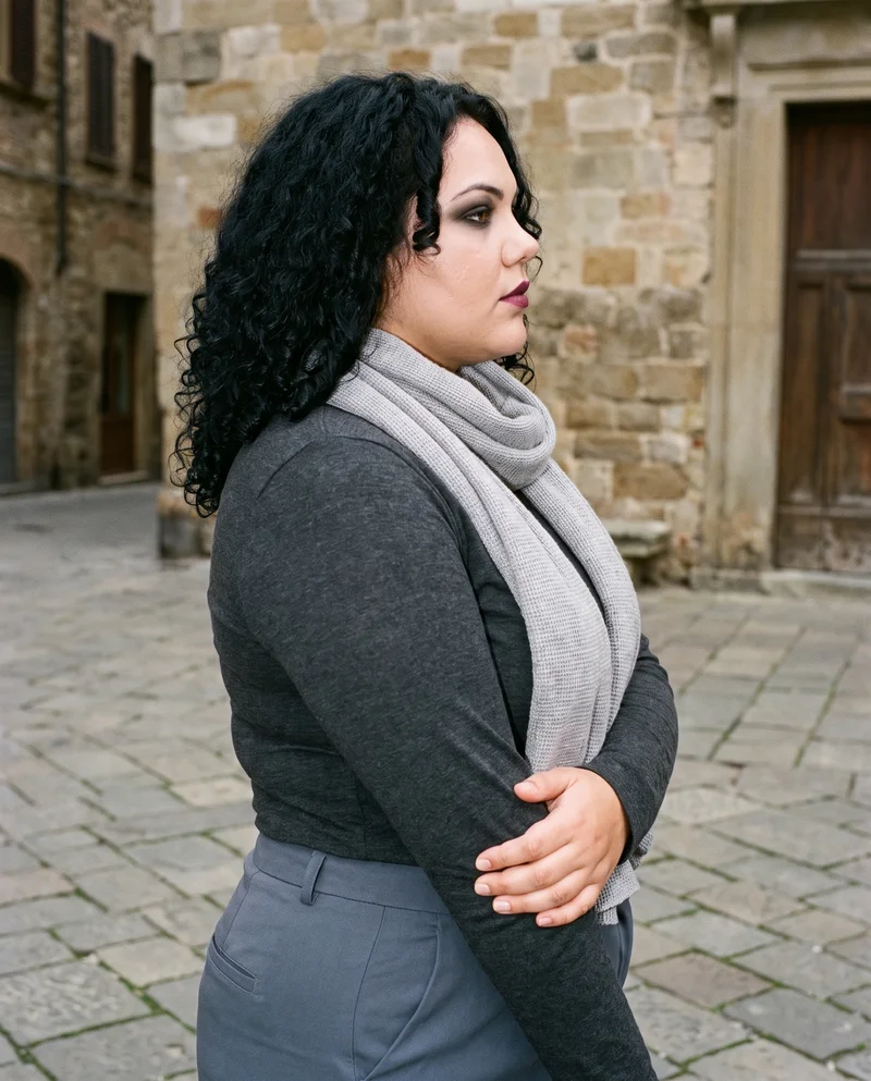 A 22-year-old Caucasian woman is shown from the upper body in side view, wearing a grey wool scarf and holding her elbow with a serious expression. A town square forms the backdrop.