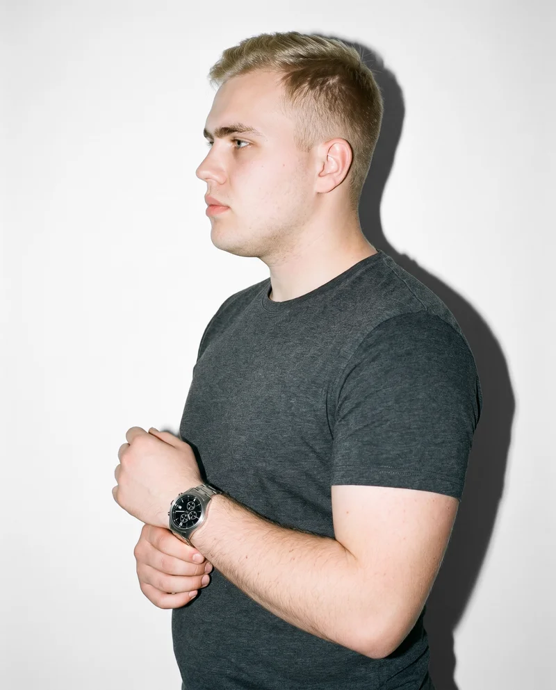 A 23-year-old Slavic man is shown in an upper-body side view, touching the silver chronograph sports watch on his wrist. The natural pose is set against a pure white studio-wall backdrop.