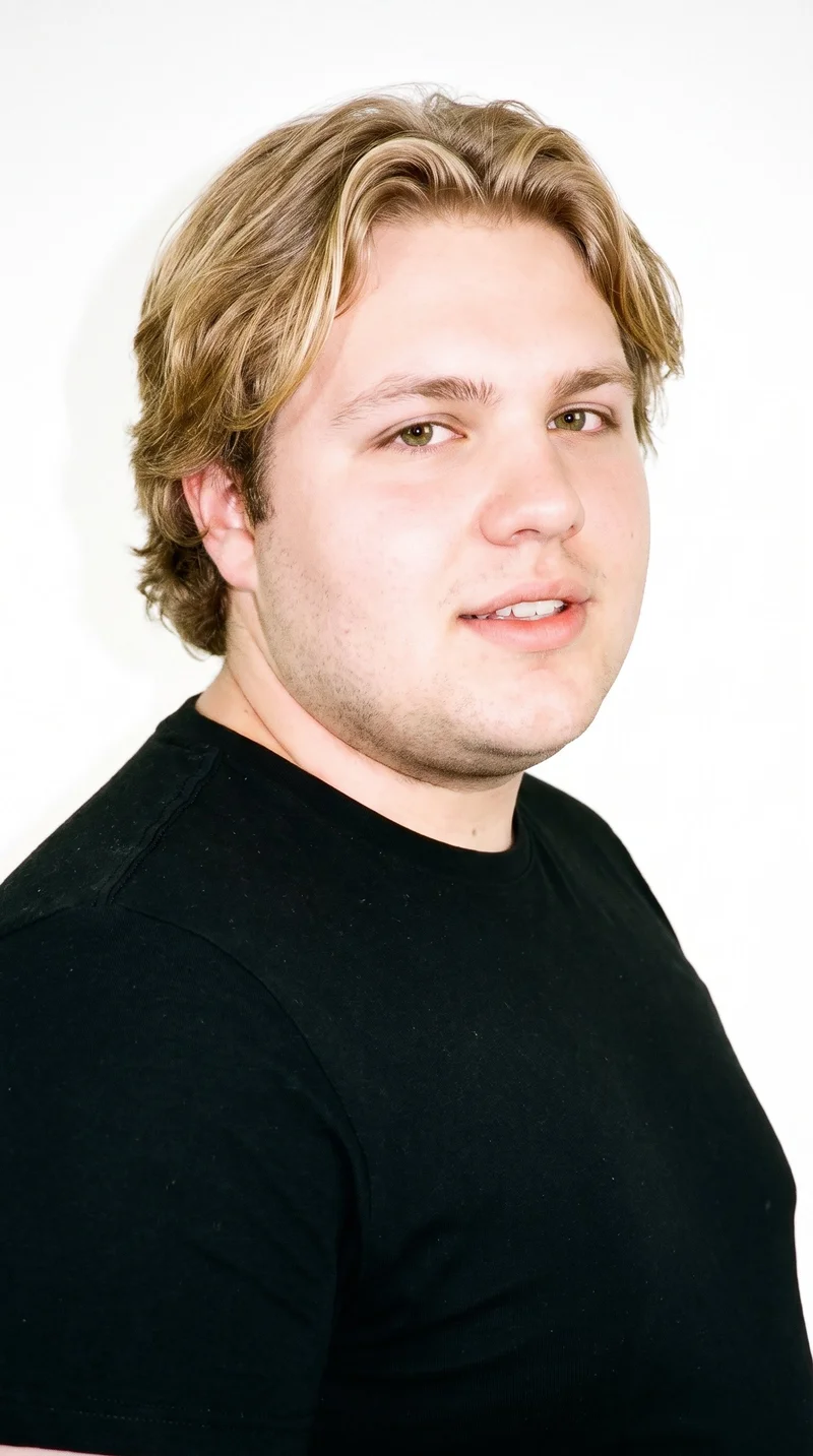 A 21-year-old Caucasian man with very pale, neutral-toned skin and a round face defined by soft, full contours and few angles. He has thin eyebrows, hazel eyes with green-gold flecks, and medium-sized lips. His mid-tone blonde hair falls in soft waves, and he is clean-shaven with a smooth face.