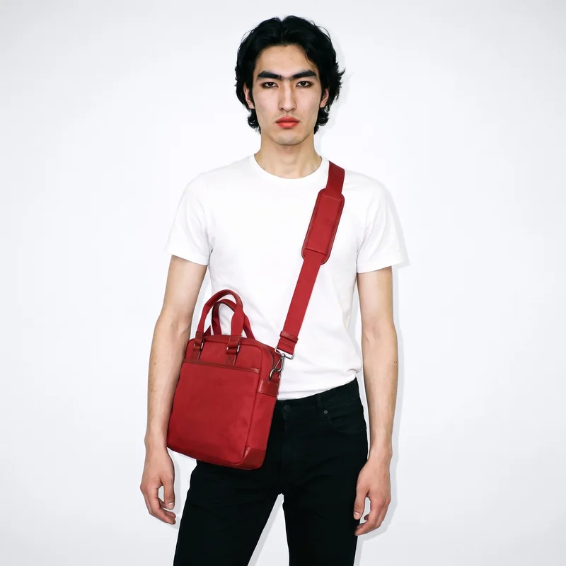 A 20-year-old Chinese man is shown from the front in a three-quarter-length view, wearing and carrying a red crossbody bag. He has a neutral expression, and the porcelain flat-color backdrop keeps the composition clean and uncluttered.