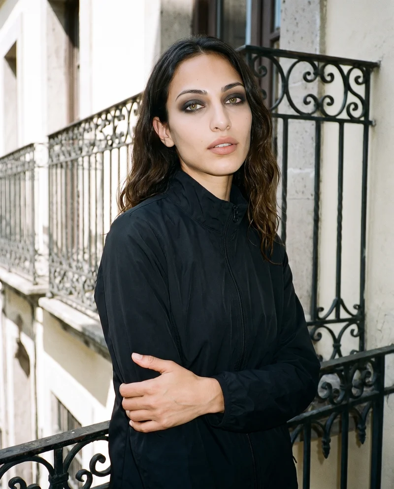 A 24-year-old Arab woman is shown from the upper body at a front three-quarter angle, wearing a black track jacket and holding her elbow. Her natural facial expression is visible against a wrought iron balcony backdrop.