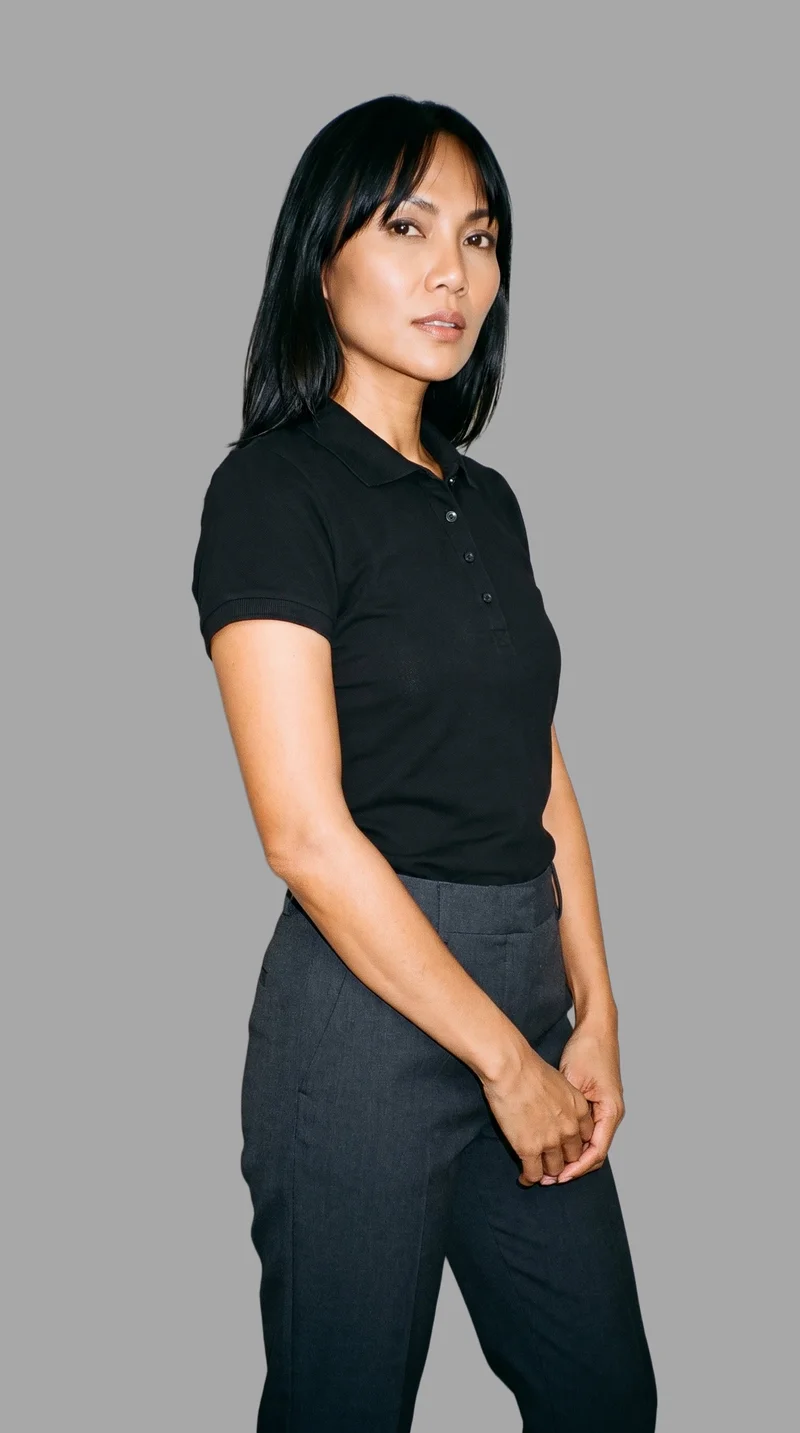 A 40-year-old Southeast Asian woman models a black women's polo shirt in a three-quarter-length, angled front view. Her hands are positioned in front, with a neutral expression, against a flat pebble-grey backdrop.