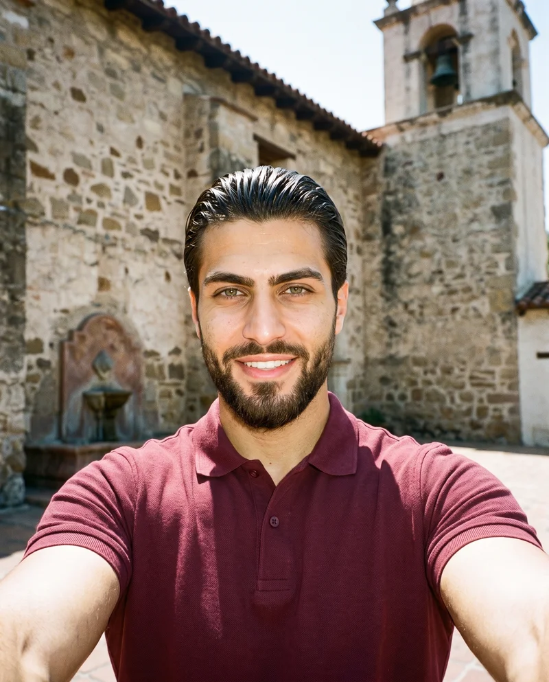 A 24-year-old Arab man wears a burgundy pique polo shirt in a front-view selfie. He is smiling with his chin down, with the Mission Courtyard Bell Fountain serving as the backdrop.