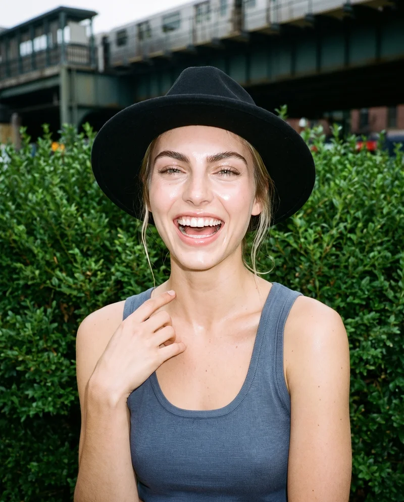A 21-year-old Caucasian woman is shown from the bust up in a front view, laughing with one hand at her collarbone while wearing a black wool fedora. An elevated park railway forms the backdrop.