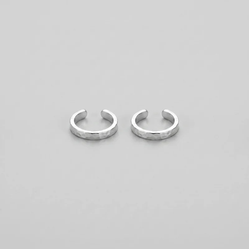 Two matching miniature silver ear cuffs are shown side by side. Each has a compact C-shaped partial-hoop profile, a narrow flattened band, a small cartilage opening, rounded ends, and subtly irregular faceted texture across the hammered outer surface.