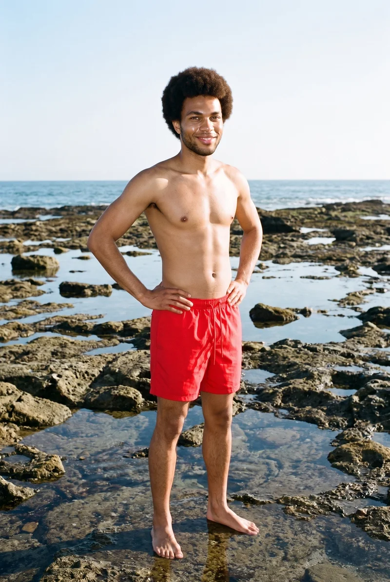 A 21-year-old African man wears red swim trunks in a full-body, angled-front view. He stands with his hands on his hips and a smile, against a backdrop of tidal pool rocks.