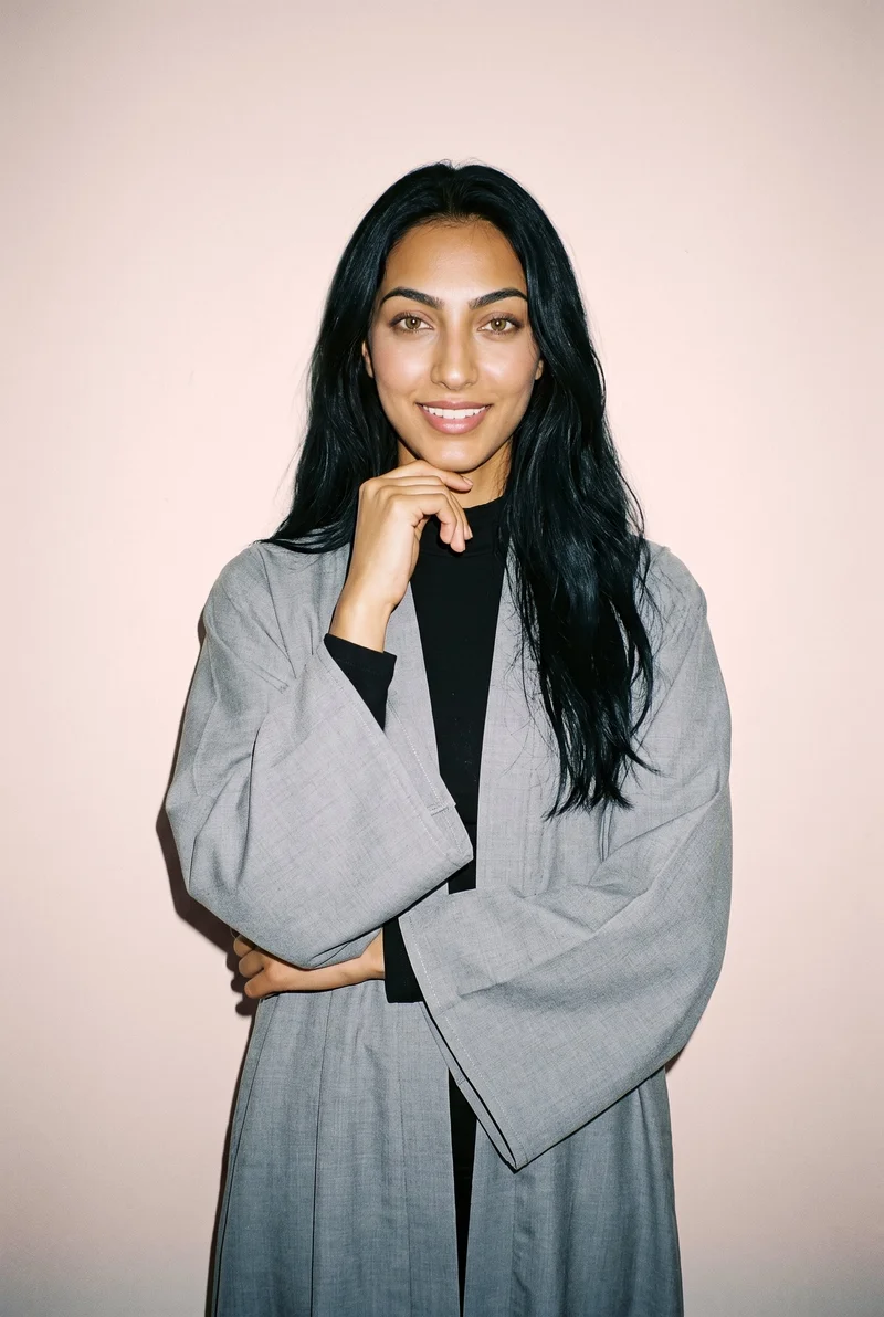 A smiling 19-year-old Arab woman wears a grey kimono abaya in an upper-body, front-facing view, with one wrist held at her jaw. A powder-pink studio-wall backdrop frames the composition.