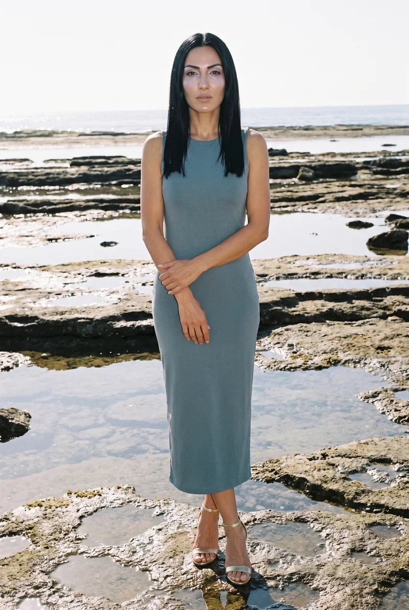 A 42-year-old Turkish woman is shown full body from the front wearing women's gold heeled sandals. Her arms are held low and her facial expression is neutral, with a tidal pool rocks backdrop.