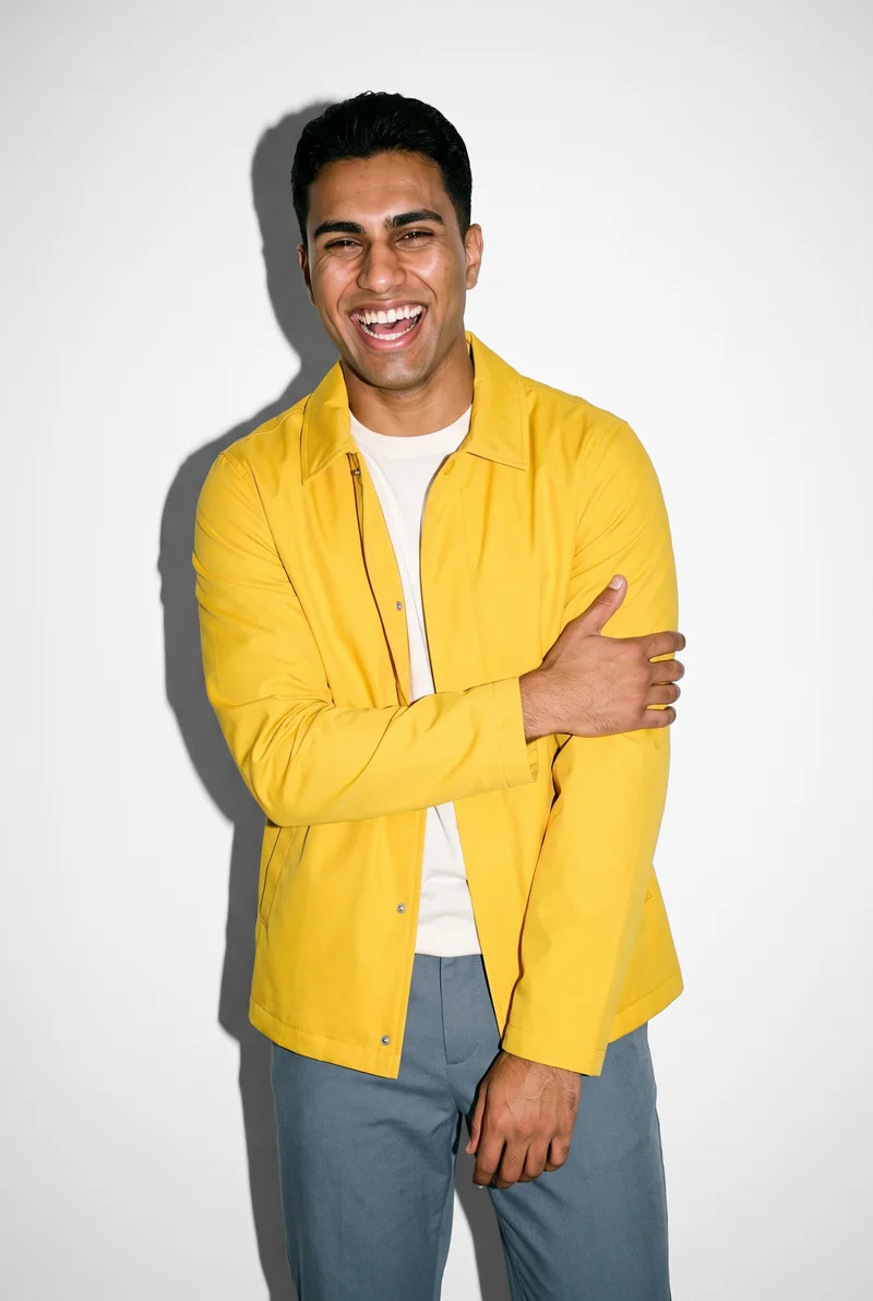 A 19-year-old Arab man is shown from the front in a three-quarter-length view, laughing while holding one arm, and wearing a yellow rain mac. The garment is presented against a pure white studio-wall backdrop.