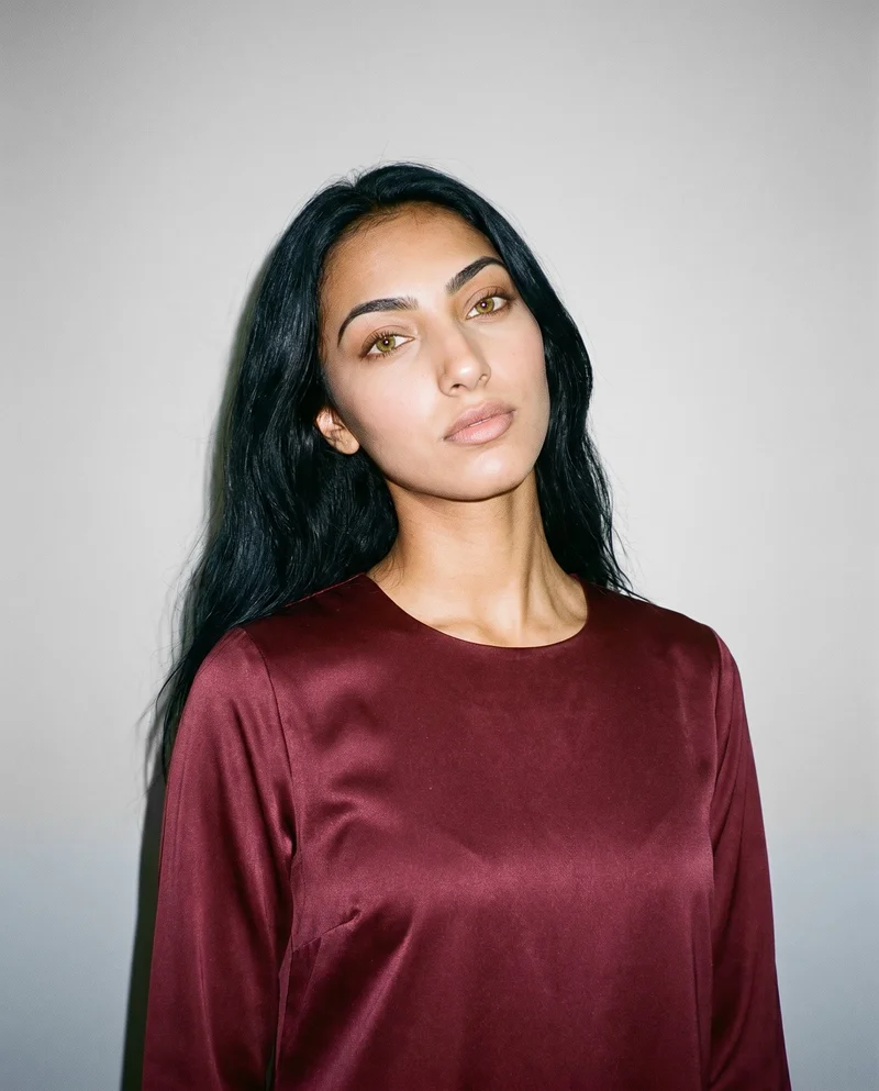 A 19-year-old Arab woman wears a burgundy satin abaya in a front-facing bust view. Her head is tilted with a neutral expression, against a pearl grey studio-wall backdrop.