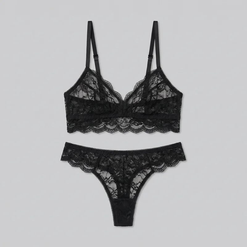 A matching black lace bralette and thong are arranged in a mostly complete front-facing flat lay. The bralette features triangular unpadded cups, a deep V plunge neckline, narrow adjustable shoulder straps with visible sliders, a wide underbust band, and scalloped edging. The low-rise thong has a narrow front panel, high-cut leg openings, and scalloped trim along the waist and leg edges, with sheer floral lace throughout.