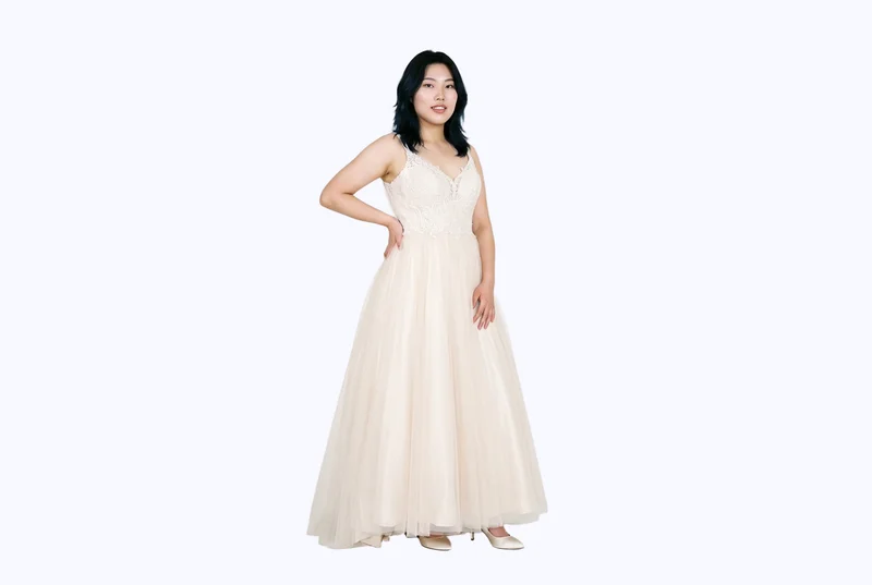 A 22-year-old Chinese woman wears a champagne ballgown wedding dress in a full-body front three-quarter view, standing with one hand on her hip and a curious expression. The dress is shown against a flat porcelain-colored backdrop.