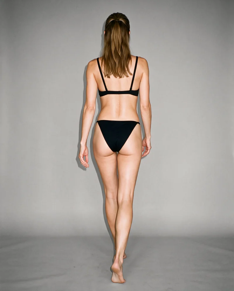 A 36-year-old Slavic woman is shown full body from the rear while walking, wearing a black triangle bikini. A flat Couture Grey backdrop fills the background.