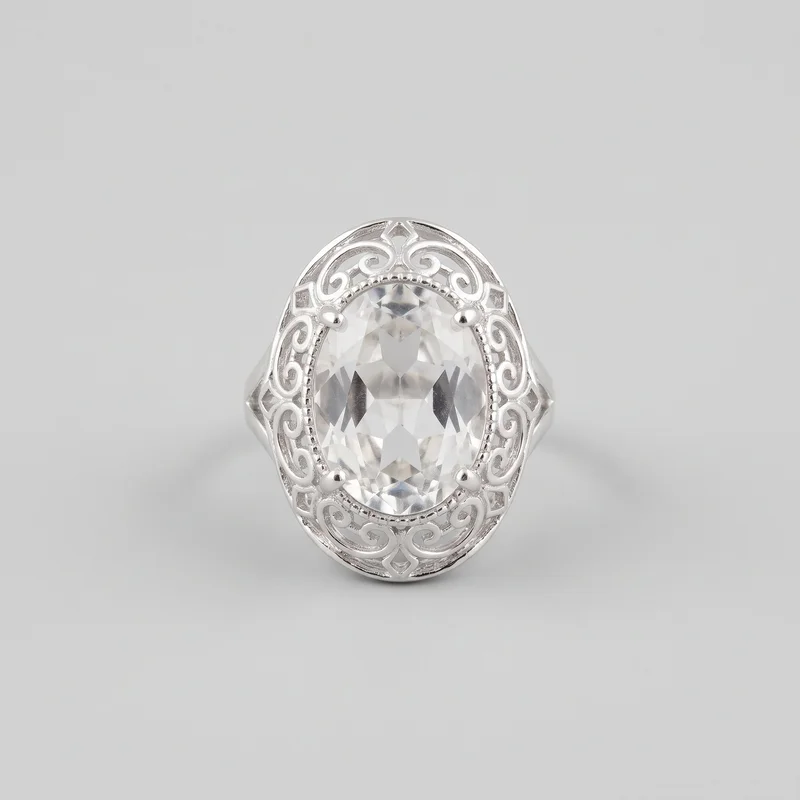 A silver cocktail ring faces forward in a close standalone view. Its vertically oriented oval faceted center stone is held by four rounded prongs and framed by a raised openwork oval surround with symmetrical scroll filigree, bead-like inner edging, and diamond-shaped accents at the top, bottom, and sides. Open split shoulders taper into the partial band.