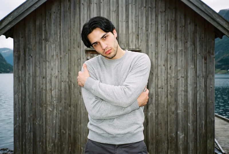 An 18-year-old Slavic male model wears a grey crewneck sweatshirt in an upper-body, front-view composition. He poses with his arms crossed in a self-hug and has a confident facial expression. A Norwegian fjord village appears as the backdrop.