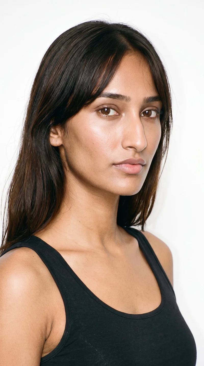 A 20-year-old Indian woman with medium-brown skin and a neutral undertone. Her face is longer than it is wide, with a tall forehead and elongated cheeks. She has thick eyebrows, medium lips, and very dark, near-black eyes. Dark-brown hair falls in longer curtain bangs with a centre part, framing both sides of her face.