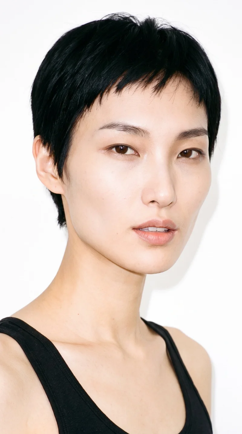 This 46-year-old Korean woman has light porcelain skin with a warm yellow undertone and an angular face defined by sharp cheekbones and jawline. She has thin eyebrows, brown eyes, and medium-sized lips. Her deep black hair is styled in a very short pixie cut, close at the sides with more length on top.
