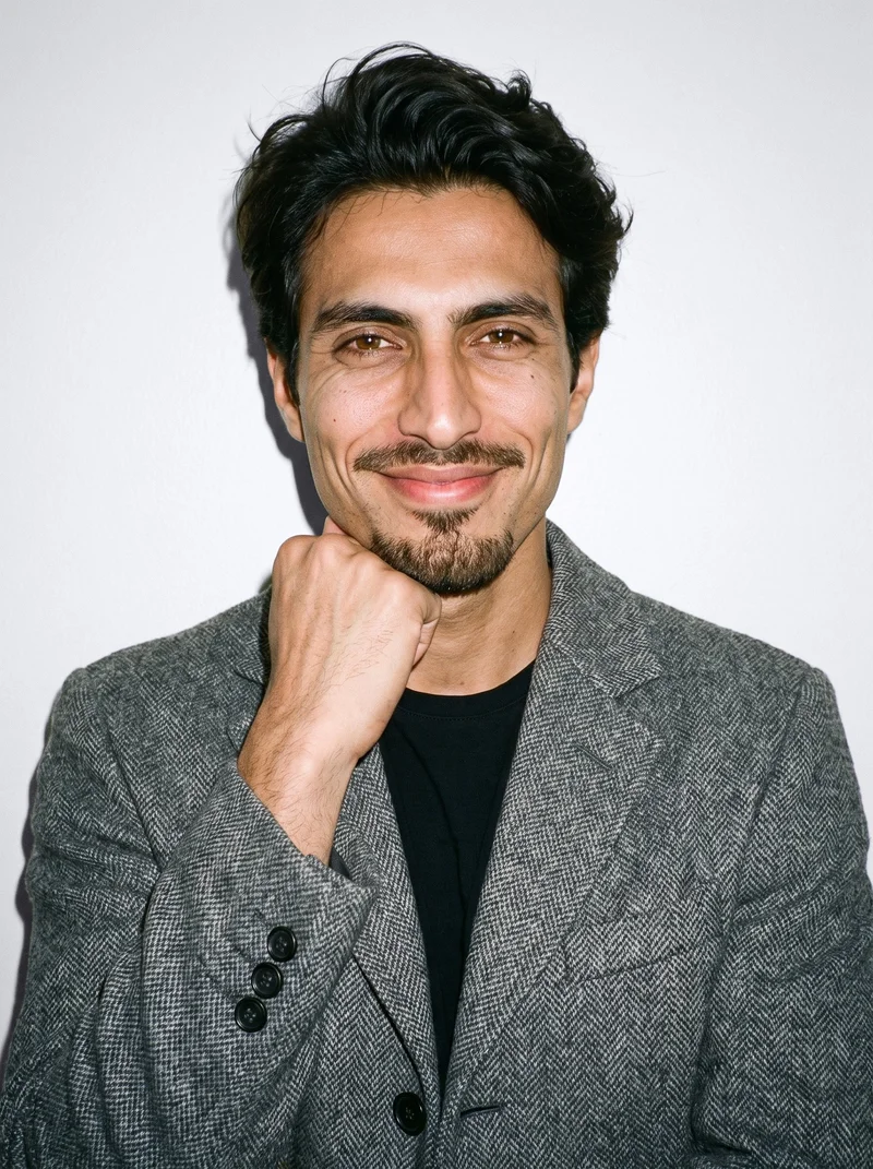 A 34-year-old Arab man is shown in a front-facing bust view wearing a grey wool coat, with one wrist raised at his jaw and a smiling expression. A porcelain studio-wall backdrop frames the composition.